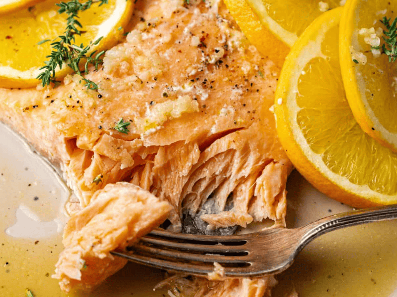 Cooked salmon fillet garnished with lemon slices and herbs, with a fork flaking the fish on a plate with sauce.