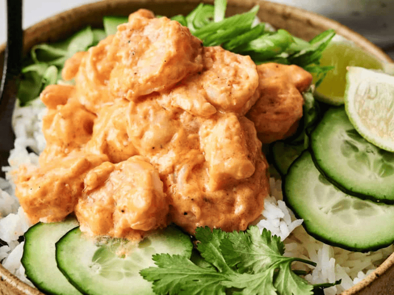 A bowl of rice topped with shrimp in a creamy sauce, garnished with cucumber slices, lime wedge, cilantro, and sliced green onions.