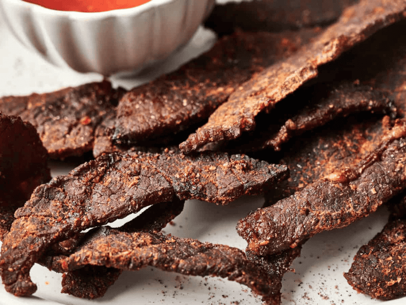 A plate of beef jerky slices next to a small bowl of dipping sauce.