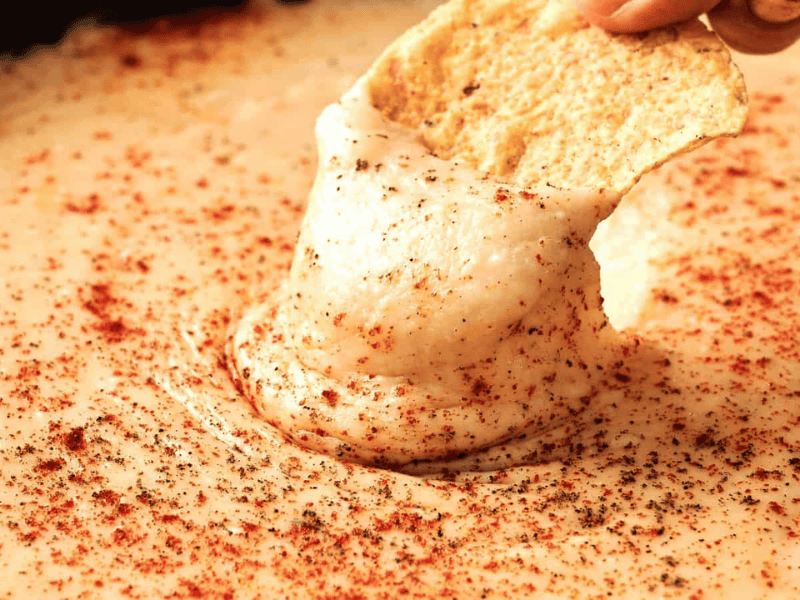 A hand dips a tortilla chip into creamy cheese dip sprinkled with paprika and black pepper.