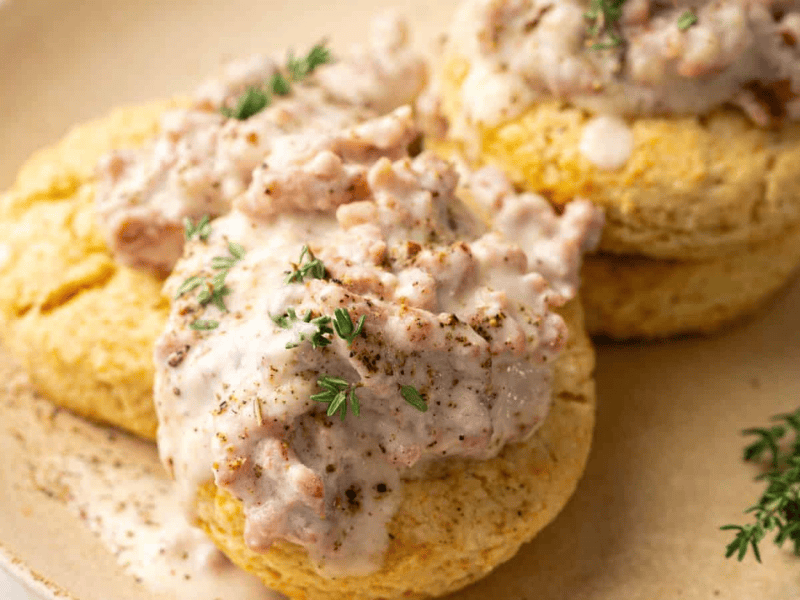 Two biscuits topped with creamy sausage gravy and garnished with fresh thyme are served on a beige plate.