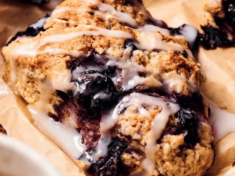 A close-up of a blueberry scone with a light glaze, resting on brown parchment paper.