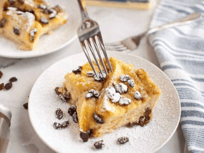 A fork cutting into a slice of bread pudding with raisins, dusted with powdered sugar, served on a white plate.