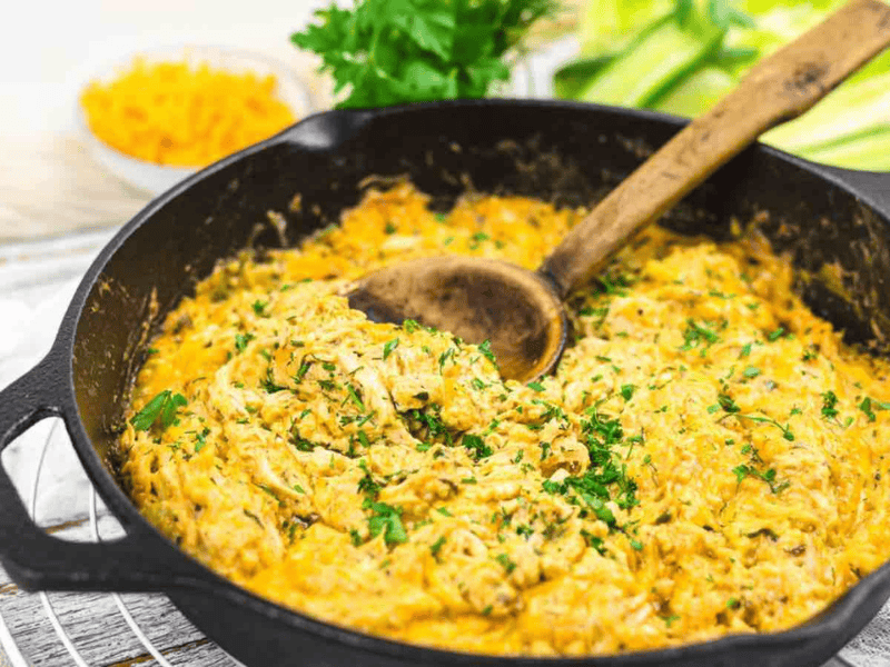 A cast iron skillet filled with a creamy dish, garnished with chopped herbs and a wooden spoon inside. Fresh parsley and sliced vegetables are seen in the background.
