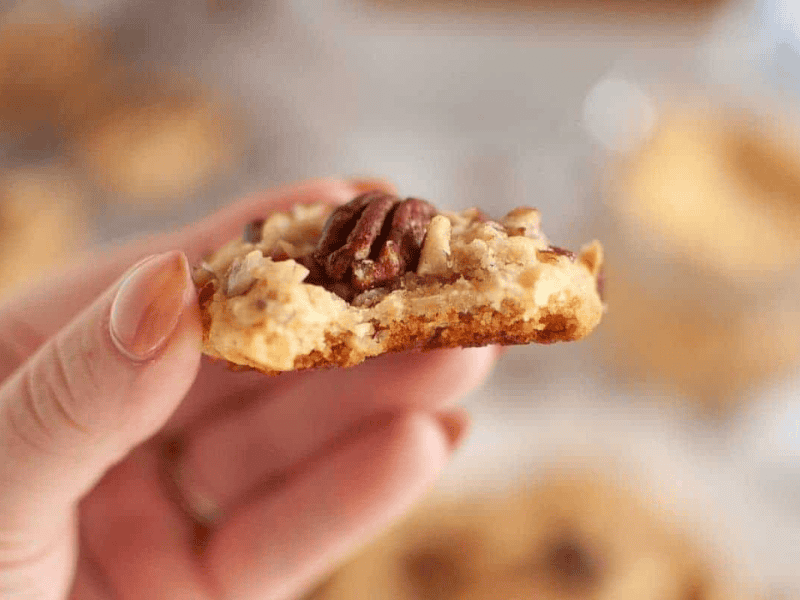 A hand holds a bitten Butter Pecan Cookie topped with a pecan and chopped nuts, with a blurred background.