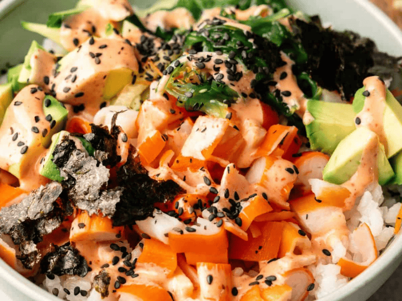 A bowl of sushi rice topped with diced avocado, raw fish, seaweed, carrots, black sesame seeds, and a creamy orange sauce.