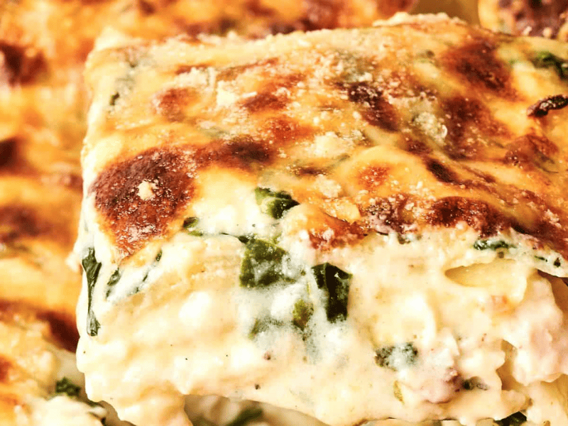 Close-up of a slice of creamy lasagna with browned cheese on top and visible layers of pasta, spinach, and sauce.