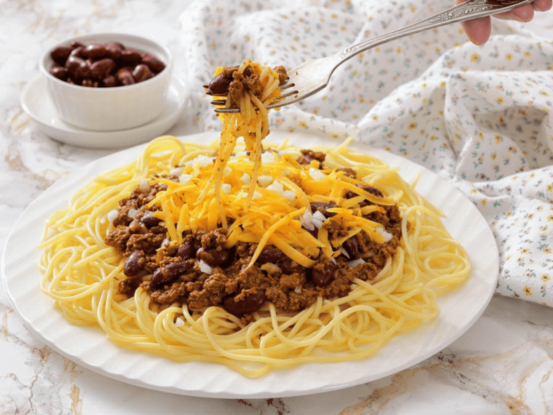 A plate of spaghetti topped with Cincinnati chili, shredded cheddar cheese, and chopped onions, with a fork lifting a bite.