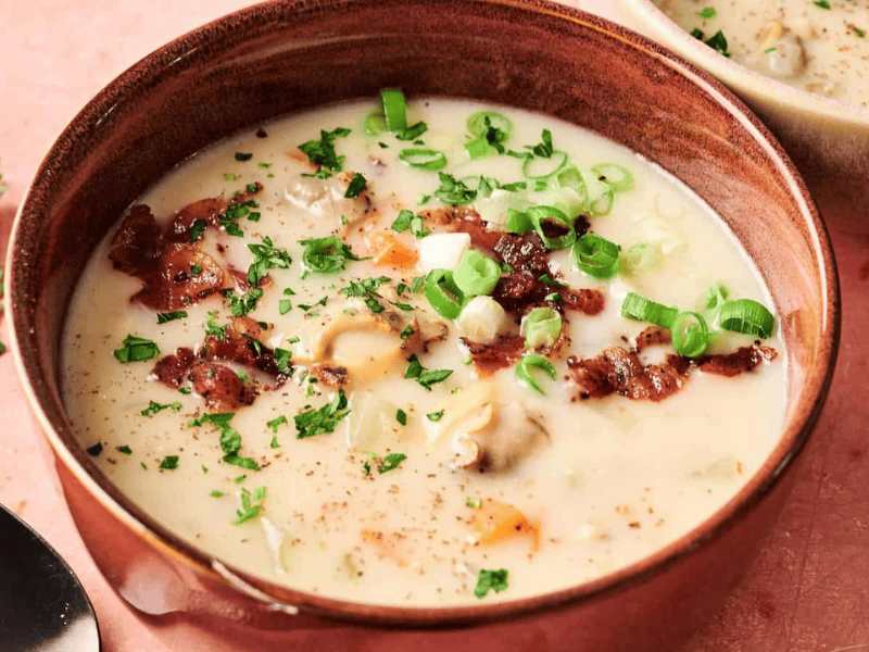 A bowl of creamy soup with clams, bacon, chopped green onions, and parsley, placed on a pink surface next to a spoon and some fresh herbs.