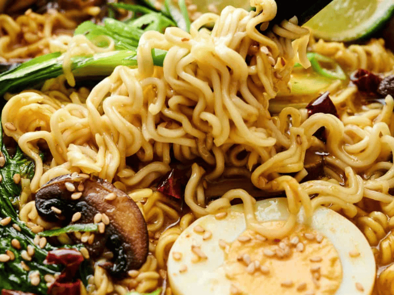 Close-up of a bowl of ramen with noodles, bok choy, mushrooms, half a boiled egg, sesame seeds, and lime, garnished with chili slices. Black chopsticks hold some noodles above the bowl.