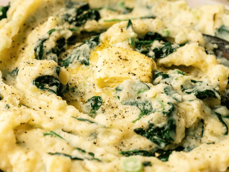 A bowl of mashed potatoes with spinach, topped with a pat of butter and black pepper, with a wooden spoon partially visible.