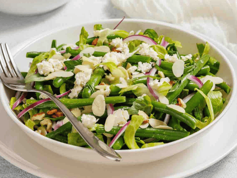 A bowl of green bean salad with feta cheese, sliced almonds, red onions, and lettuce, served with a fork and spoon.