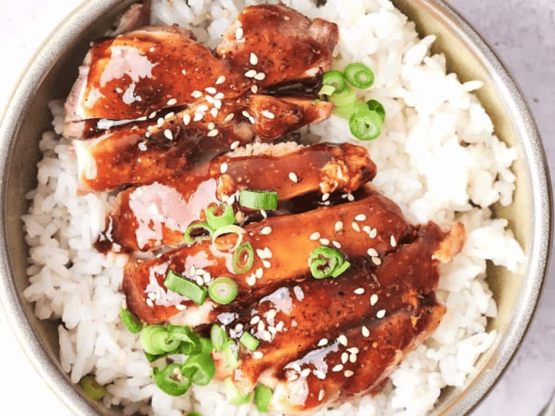 A bowl of white rice topped with glazed chicken and garnished with green onions and sesame seeds.