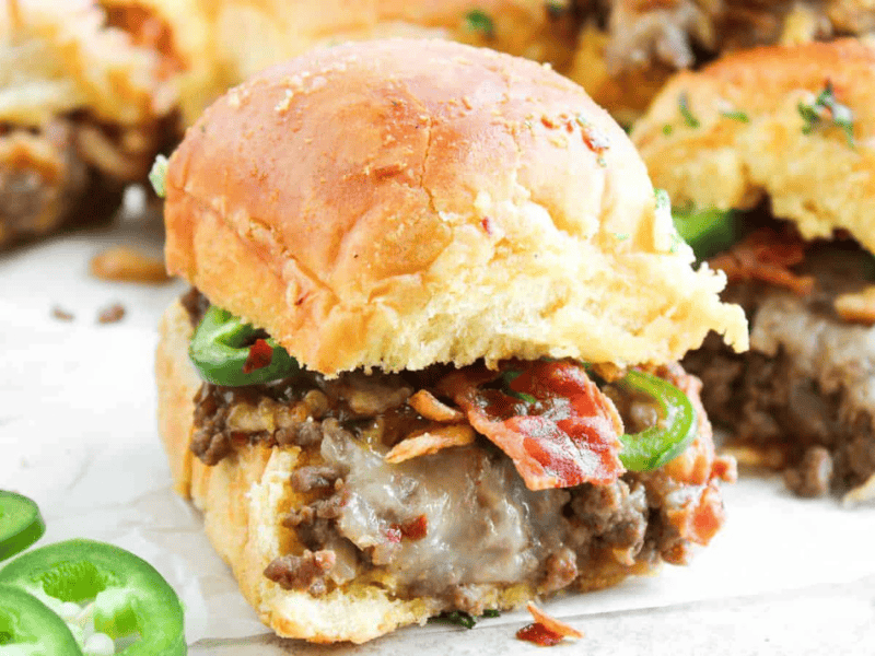A close-up of sliders filled with cooked ground beef, melted cheese, bacon, and sliced jalape&ntilde;os on a white surface, with some jalape&ntilde;o slices scattered nearby.