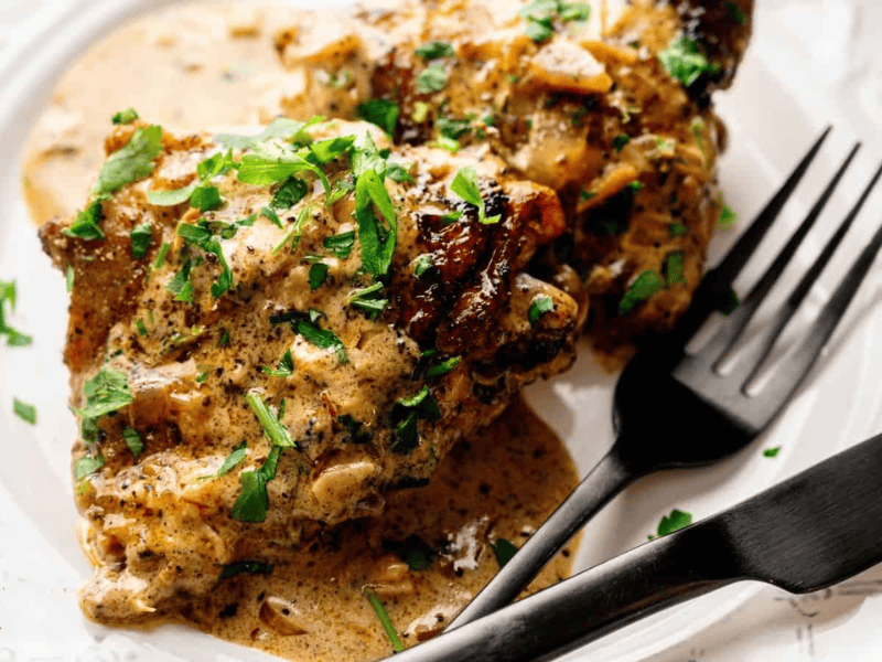 Two pieces of seasoned, creamy chicken garnished with chopped herbs on a white plate, with a black fork and knife beside them.