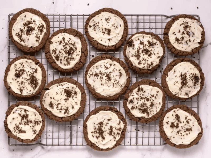A cooling rack with twelve Crumbl Copycat Frosted Oreo Cookies topped with white frosting and chocolate crumbs, arranged on a white marble surface.