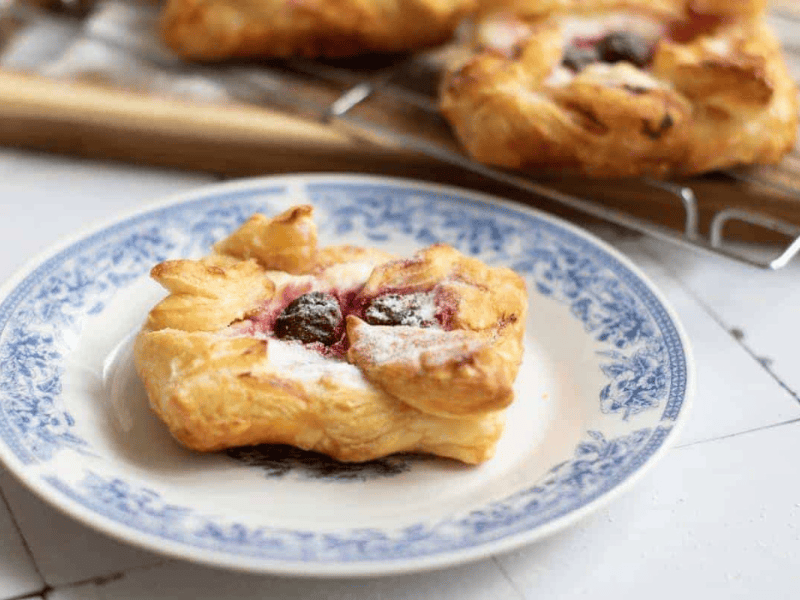 A cherry Danish pastry with powdered sugar sits on a white plate with a blue floral pattern; more pastries are on a cooling rack in the background.