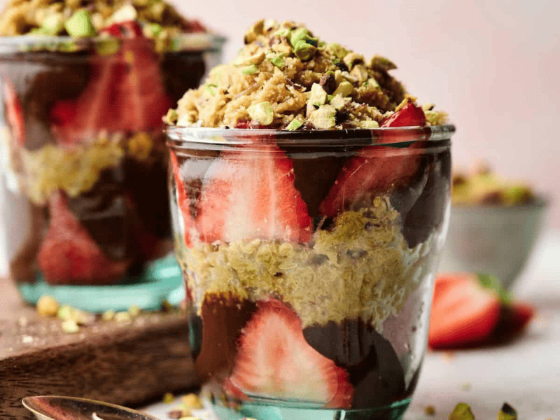 Two glass cups filled with layers of sliced strawberries, chocolate, granola, and chopped pistachios, with a blurred bowl of nuts in the background.