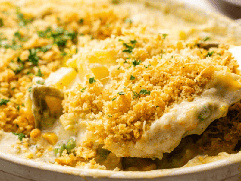 A spoon scoops a portion of creamy vegetable casserole topped with golden breadcrumbs and chopped herbs.