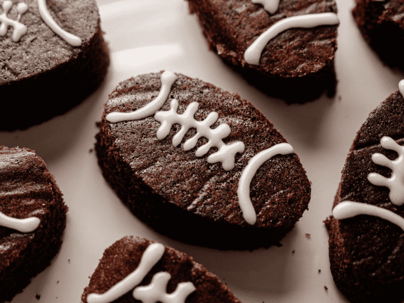 Oval-shaped brownies decorated with white icing to resemble footballs, arranged on a white surface.
