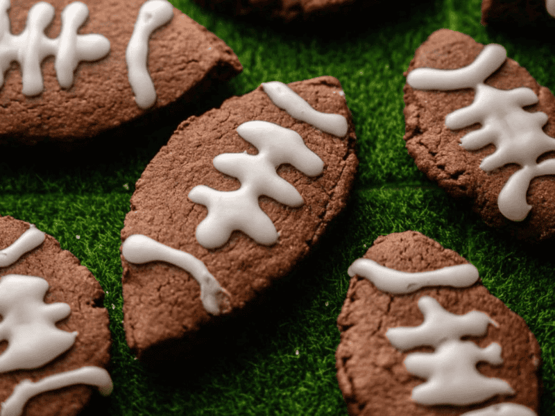 Football-shaped chocolate cookies with white icing laces are arranged on a green textured surface resembling grass.