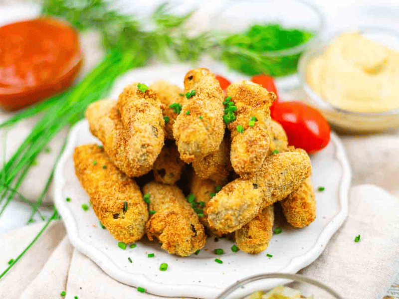 A plate of breaded and baked pickles garnished with chopped chives, surrounded by fresh herbs, grape tomatoes, and dipping sauces.