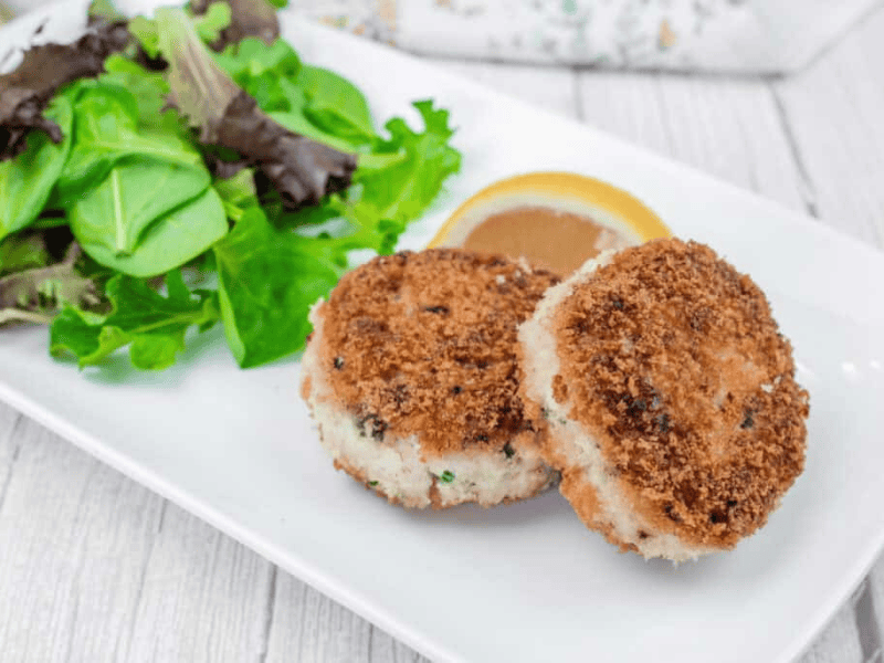 Two Salmon Cakes on a white plate with mixed leafy greens and a lemon wedge.