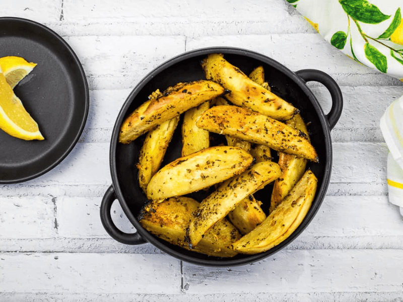 Round black dish filled with seasoned roasted potato wedges, placed on a white wooden surface alongside a white and yellow striped cloth napkin.