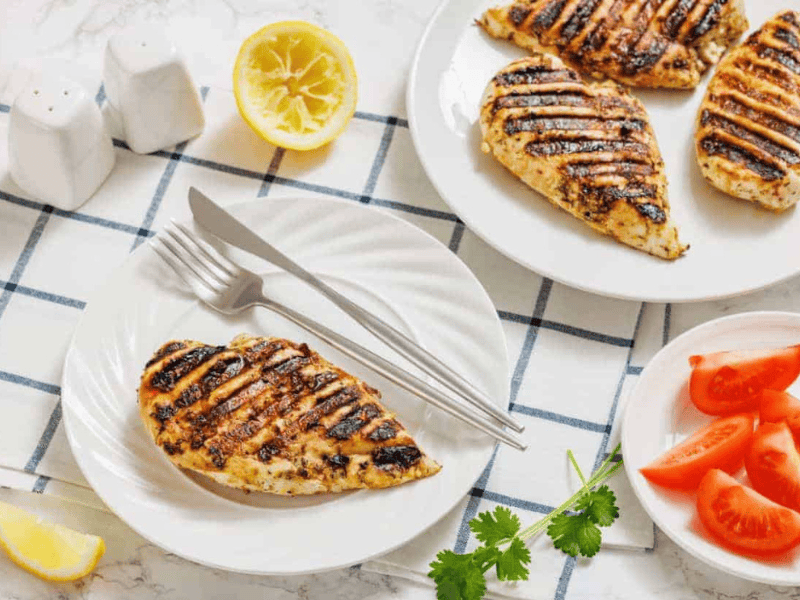 Grilled chicken breast on a white plate with fork and knife, sliced tomatoes, lemon halves, cilantro, and salt and pepper shakers on a checkered cloth.