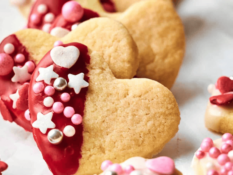 Heart-shaped cookies partially dipped in red icing, decorated with pink, white, and silver sprinkles, including stars and hearts, scattered on a white surface.
