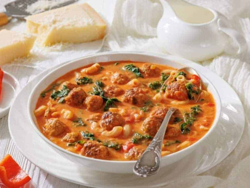 A bowl of meatball soup with pasta, spinach, and tomato broth, served with a spoon.