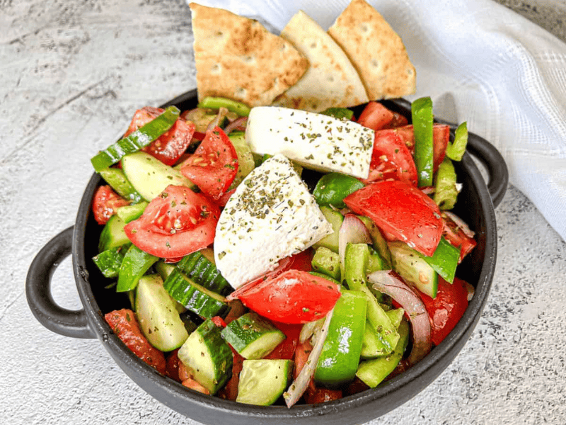 A black bowl contains a Horiatiki salad with blocks of feta cheese, garnished with herbs.
