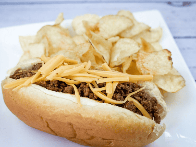 A hot dog chili topped with shredded cheddar, served with potato chips on a white plate.