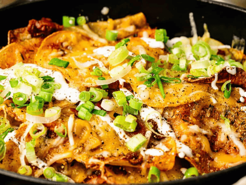 A close-up of a skillet filled with cheesy nachos topped with sliced green onions, melted cheese, sour cream, and various seasonings.