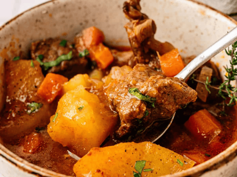 A bowl of beef stew with chunks of beef, potatoes, carrots, and herbs in a rich broth, with a spoon inside the bowl.