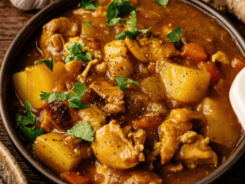 A bowl of chicken stew with chunks of potato and carrot, garnished with chopped cilantro, on a wooden surface.