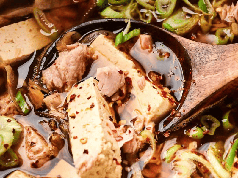 Close-up of a wooden spoon in a vibrant kimchi stew with tofu and pork, garnished with sliced green onions and red pepper flakes.