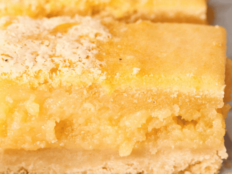 Close-up of a lemon bar with a crumbly, light yellow top layer and a thicker, moist filling resting on a shortbread crust.