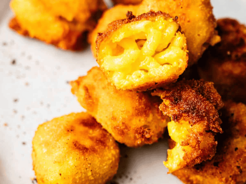 Five pieces of crispy, golden-brown fried macaroni and cheese bites are stacked on a white plate, with one bite showing the cheesy pasta inside.