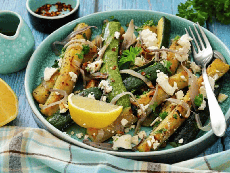 A plate of grilled zucchini with crumbled feta, sliced onions, fresh parsley, and a lemon wedge, served on a blue table with a fork and a checkered napkin.