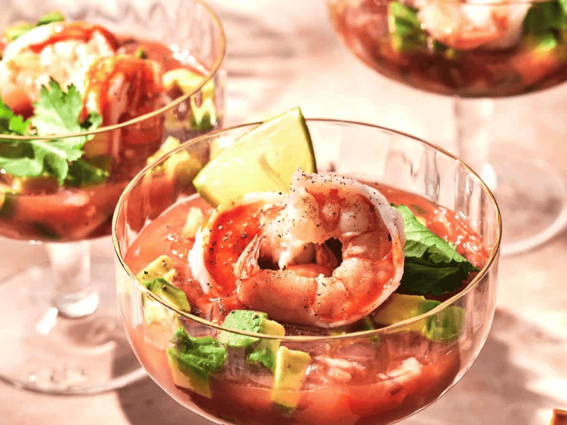 Three glasses of shrimp cocktail with avocado, lime wedge, and cilantro garnish in a tomato-based sauce, placed on a light-colored surface.