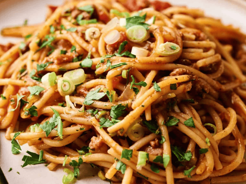 A plate of spaghetti with tomato sauce, ground meat, chopped green onions, and parsley, served with a black fork.