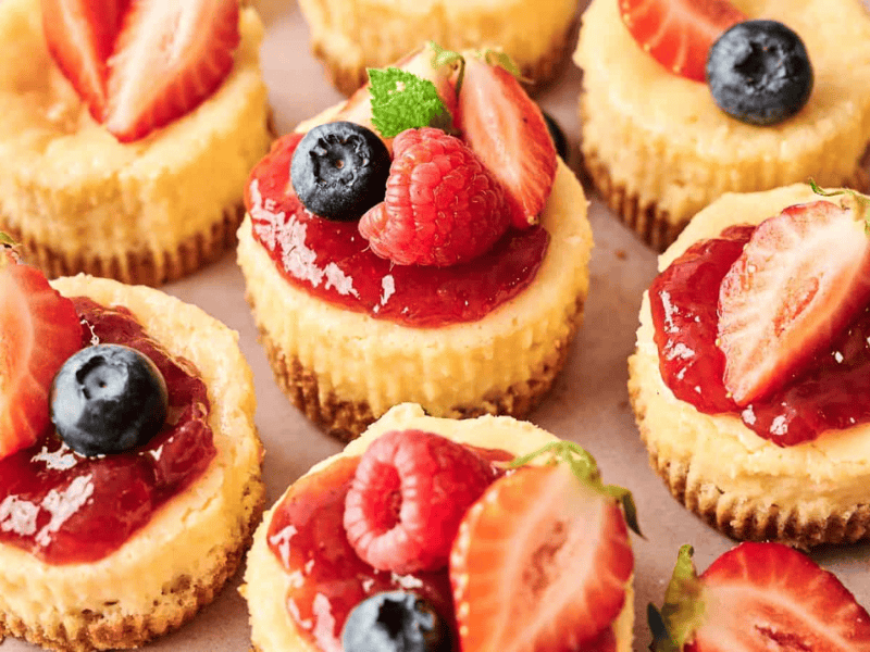 Mini cheesecakes topped with strawberry sauce, fresh strawberries, raspberries, blueberries, and a small mint leaf on a light surface.