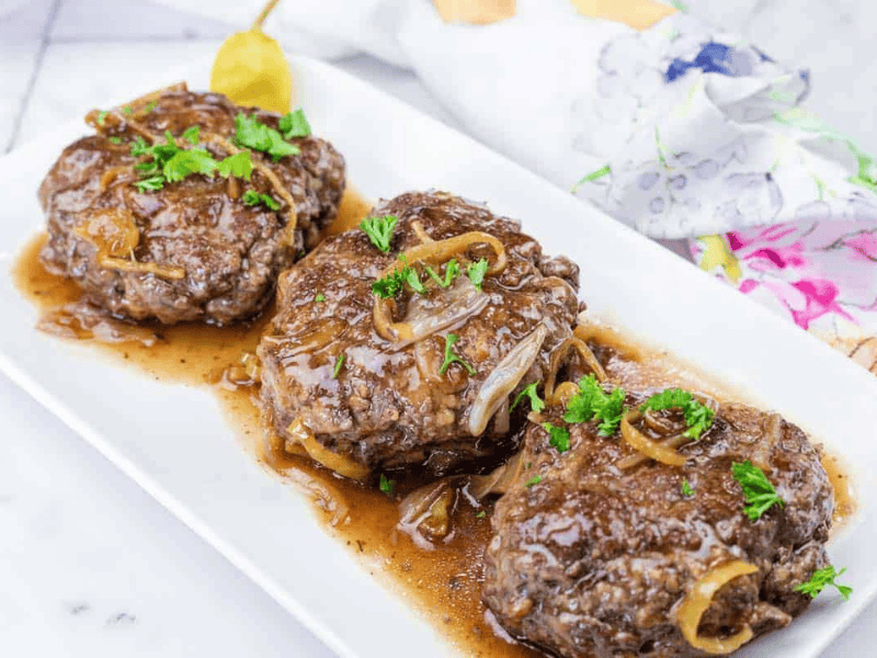 Three Salisbury steak patties topped with onions and brown gravy, garnished with parsley, served on a white rectangular plate.