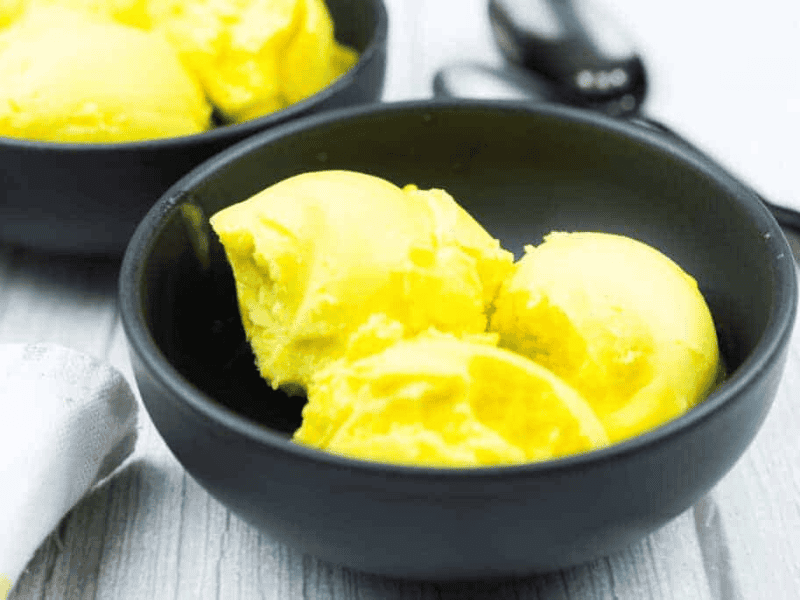 A black bowl containing three scoops of bright yellow ice cream sits on a light wooden surface, with another bowl and metal spoons in the background.