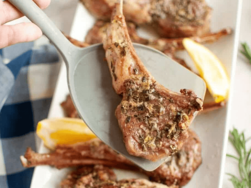 A cooked lamb chop with herbs is lifted by a spatula above a plate with more lamb chops and lemon wedges.
