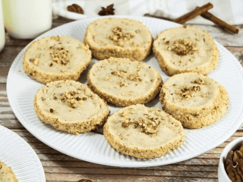 A white plate holds eight round Pecan Shortbread Cookies topped with chopped nuts, placed on a wooden surface next to bottles of milk and cinnamon sticks.