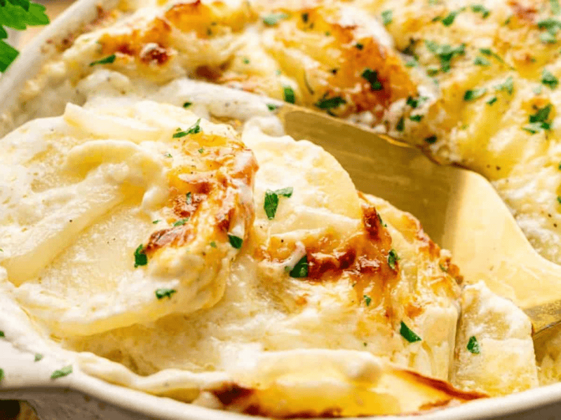 A close-up of creamy, baked scalloped potatoes with melted cheese, garnished with chopped parsley, in a white baking dish with a serving spoon.