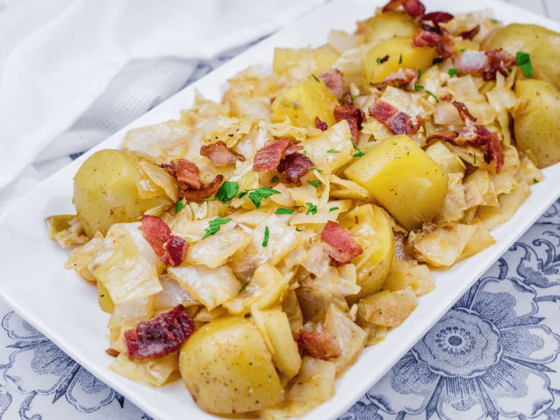 A white rectangular plate with Potatoes & Cabbage garnished with parsley on a patterned surface.