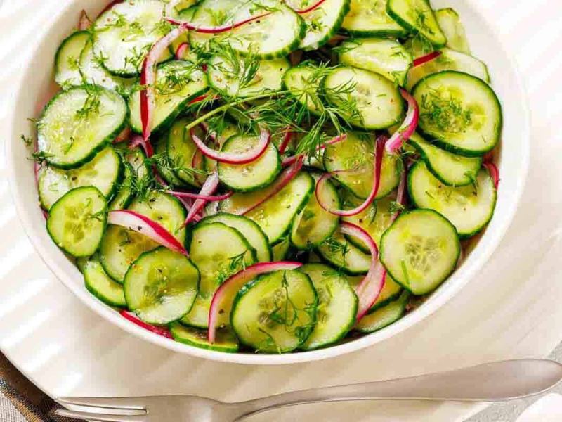A white bowl filled with sliced cucumbers, red onions, and fresh dill, placed on a white plate with a fork and spoon beside it.
